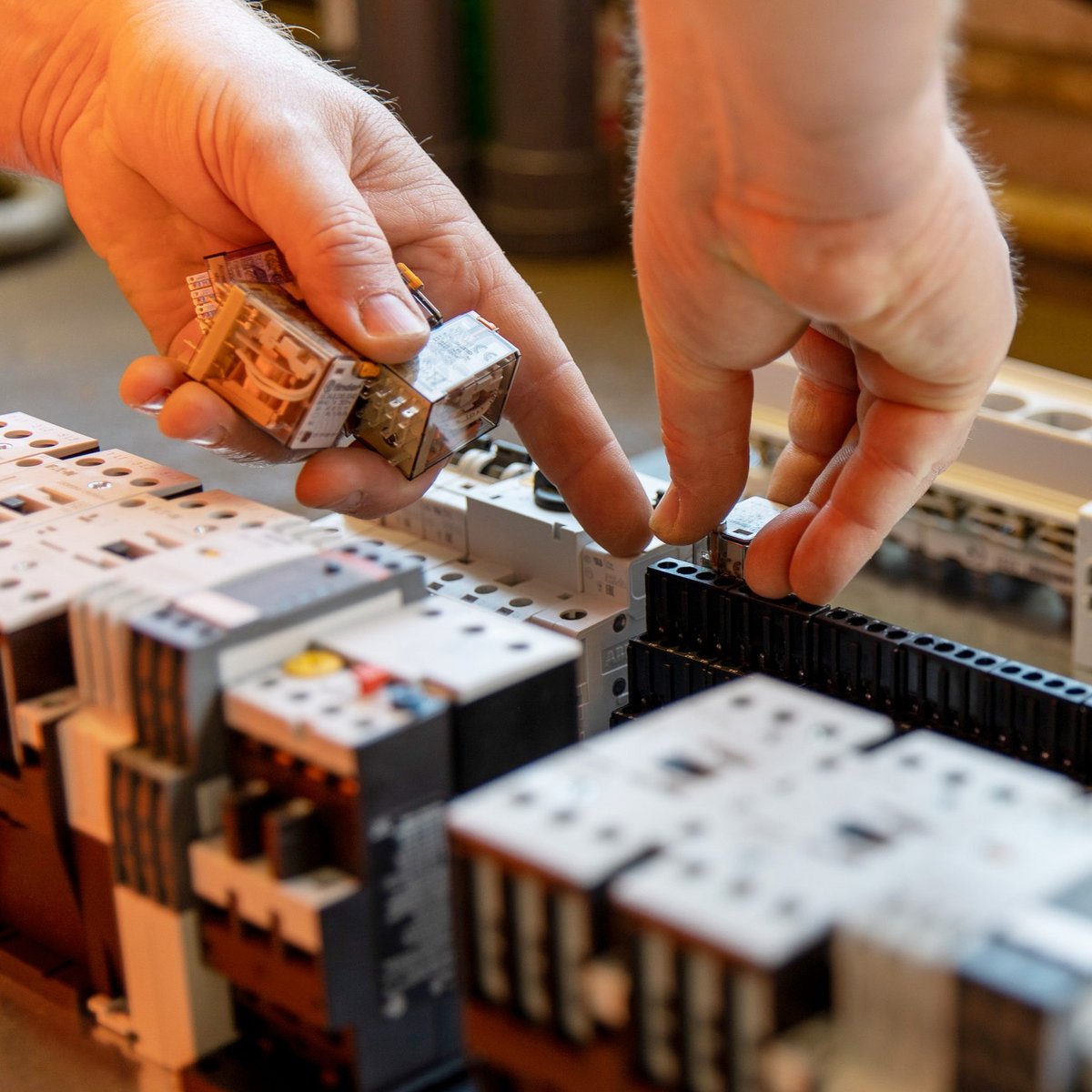 Auf einem Tisch sind mehrere elektrische Geräte angeordnet, während eine Hand ein Bauteil präzise einsetzt.