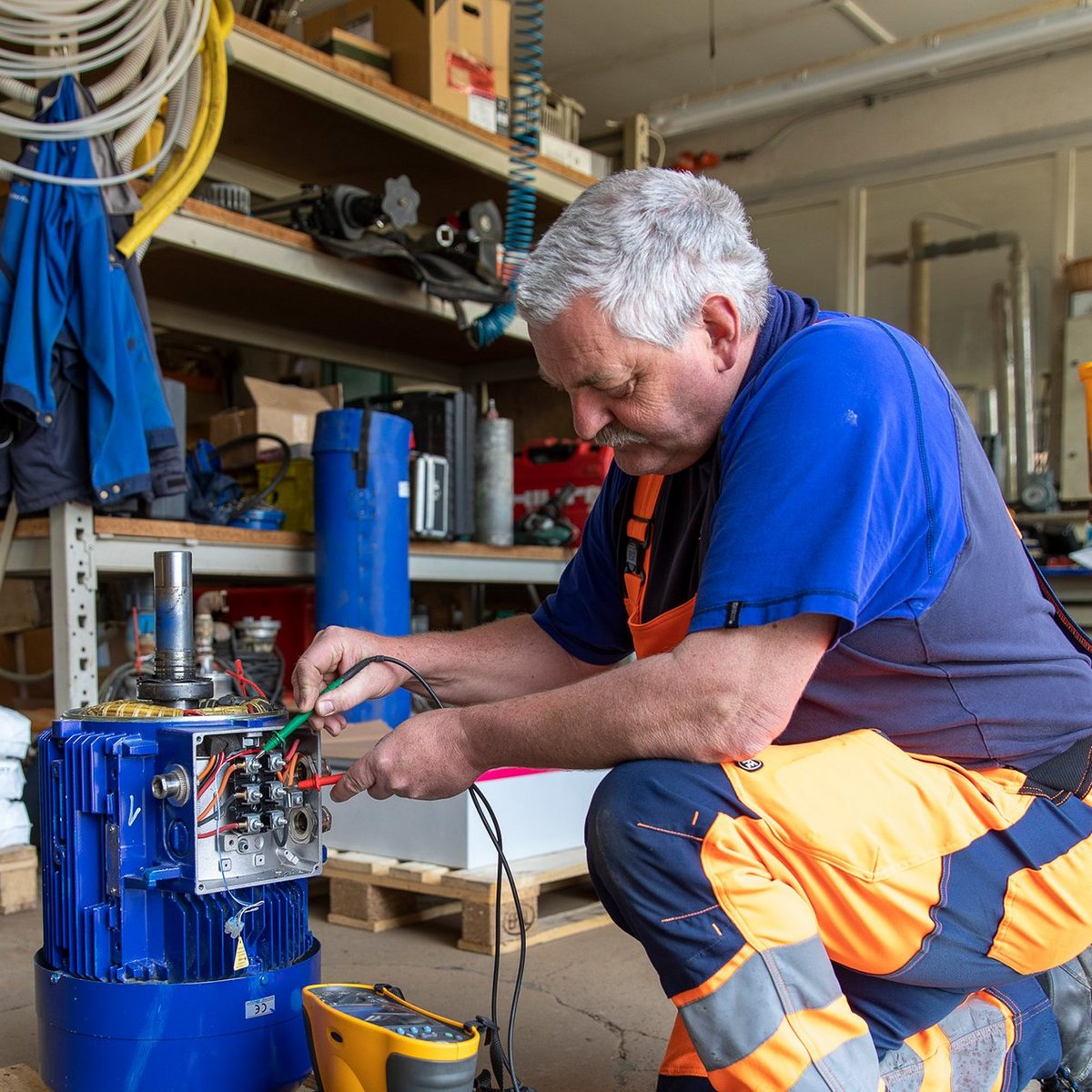 Ein Mitarbeiter der Firma Walter Fiedler in orangefarbener Arbeitskleidung repariert eine Wasserpumpe in einer Werkstatt, umgeben von Werkzeugen und Materialien.