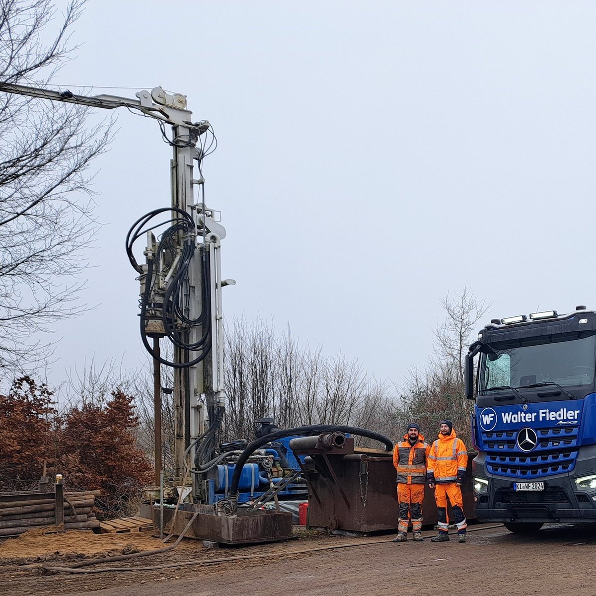 Zwei Arbeiter in orangefarbener Arbeitskleidung stehen neben einem großen Lkw und einem Servicefahrzeug auf einer Baustelle.
